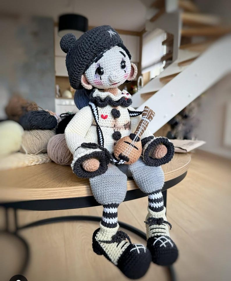 May include: A handmade crochet doll with a clown-like appearance. The doll wears a black hat, a white and black outfit, and gray pants. It holds a small ukulele. The doll's features include rosy cheeks and a whimsical expression.