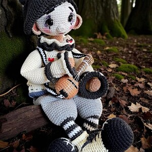 May include: A handmade crochet doll with a clown-like appearance, wearing a black hat, white face, and a ruffled collar. The doll is holding a small, brown stringed instrument and is seated in a forest setting.