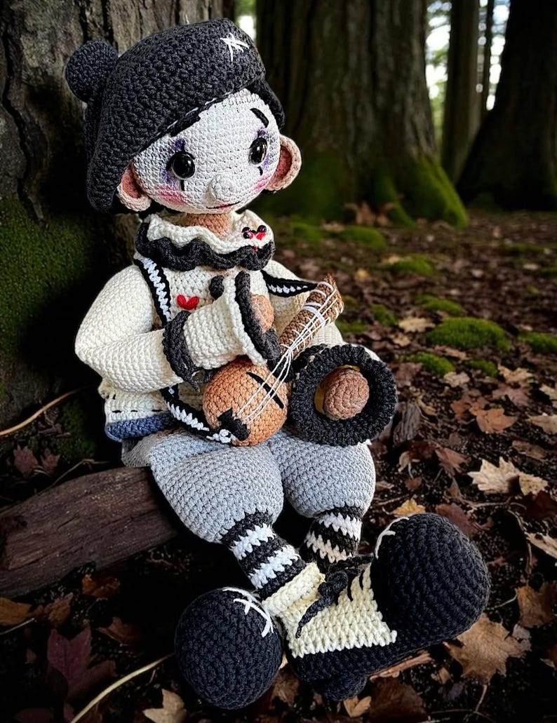 May include: A handmade crochet doll with a clown-like appearance, wearing a black hat, white face, and a ruffled collar. The doll is holding a small, brown stringed instrument and is seated in a forest setting.