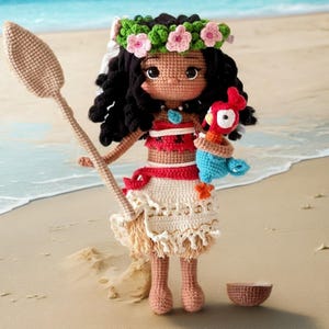 Op de afbeelding: Een gehaakte pop van Moana uit de Disney-film Vaiana, die een traditionele Hawa&iuml;aanse outfit draagt met een bloemenkrans, en een blauw-rode papegaai vasthoudt. De pop staat op een zandstrand met een halve kokosnootschelp in de buurt.