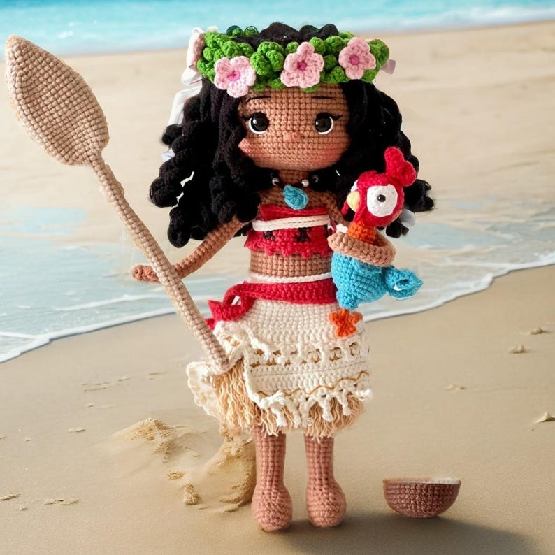 Op de afbeelding: Een gehaakte pop van Moana uit de Disney-film Vaiana, die een traditionele Hawa&iuml;aanse outfit draagt met een bloemenkrans, en een blauw-rode papegaai vasthoudt. De pop staat op een zandstrand met een halve kokosnootschelp in de buurt.