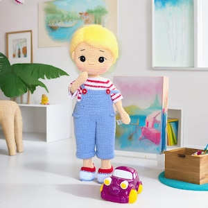 Puede incluir: Muñeca de ganchillo hecha a mano con cabello rubio, que lleva una camisa a rayas rojas y blancas y un peto azul. La muñeca está de pie junto a un coche de juguete morado con ruedas amarillas. La muñeca es una figura infantil.
