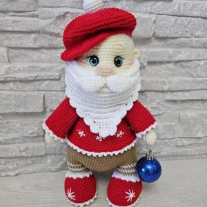 Puede incluir: Mu&ntilde;eca de ganchillo hecha a mano de Pap&aacute; Noel con gorro y chaqueta rojos, barba y ribete blancos y pantalones marrones. La mu&ntilde;eca sostiene una bola de Navidad azul. Las botas de la mu&ntilde;eca tienen detalles de copos de nieve blancos.