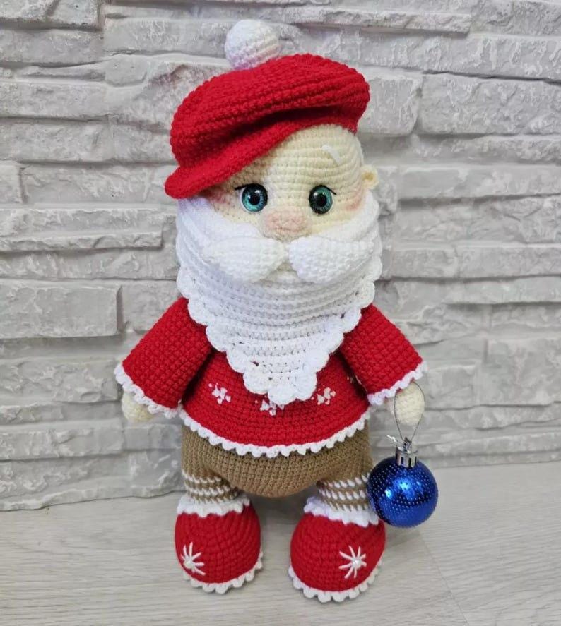 Puede incluir: Mu&ntilde;eca de ganchillo hecha a mano de Pap&aacute; Noel con gorro y chaqueta rojos, barba y ribete blancos y pantalones marrones. La mu&ntilde;eca sostiene una bola de Navidad azul. Las botas de la mu&ntilde;eca tienen detalles de copos de nieve blancos.