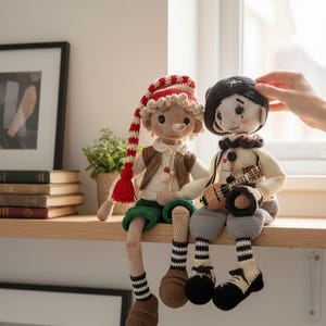 May include: Two handmade crochet dolls sit side-by-side on a wooden shelf. One doll wears a red and white striped hat, a brown vest, and green shorts. The other doll wears a black beret and holds a small ukulele. Both dolls have long legs.