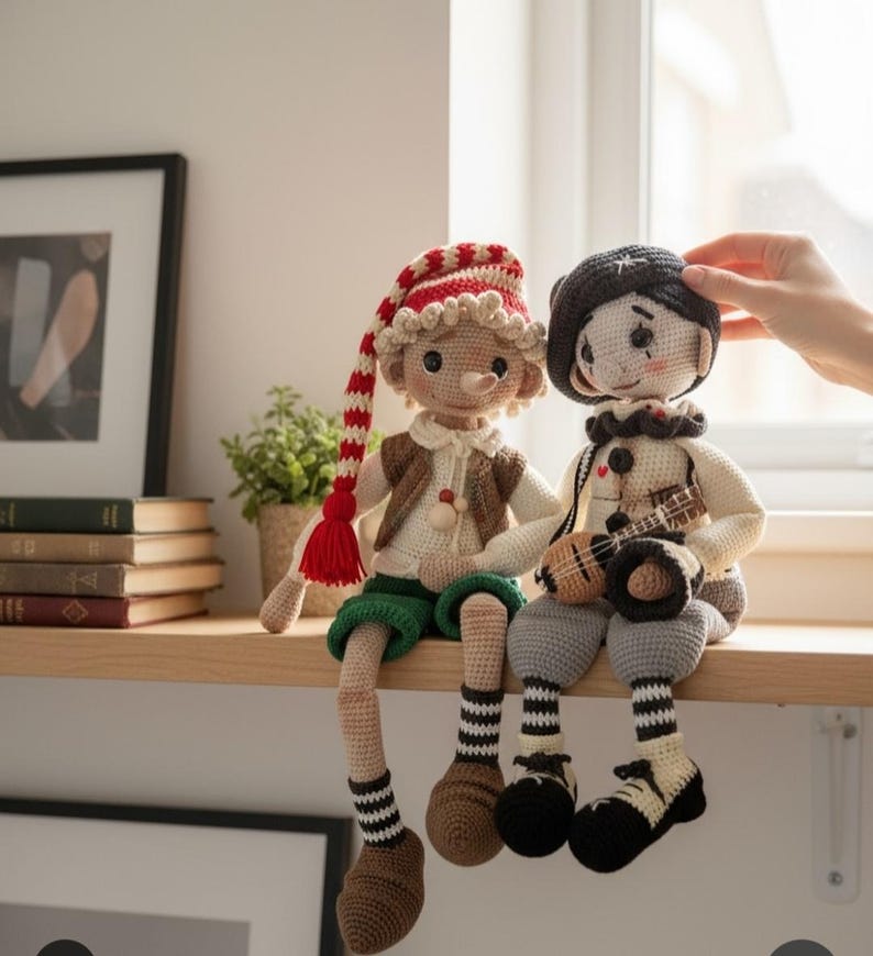 May include: Two handmade crochet dolls sit side-by-side on a wooden shelf. One doll wears a red and white striped hat, a brown vest, and green shorts. The other doll wears a black beret and holds a small ukulele. Both dolls have long legs.