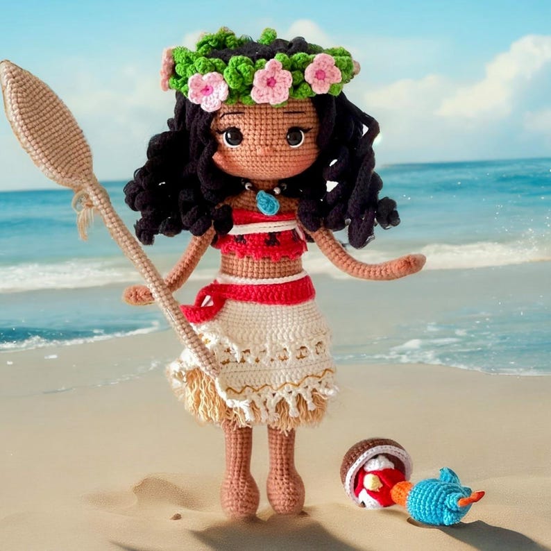 Op de afbeelding: Een gehaakte pop met een bruine huid, zwart haar en een bloemenkroon. Ze draagt een rood-witte jurk en houdt een houten peddel vast. De pop staat op een zandstrand met blauw water op de achtergrond.