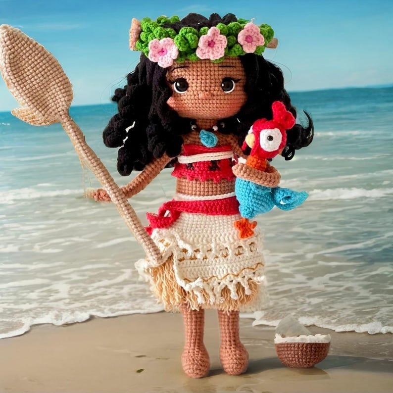 Op de afbeelding: Een gehaakte pop van Moana uit de Disney-film Vaiana, die een bruine, rode en witte outfit draagt en een blauwe en rode gehaakte papegaai vasthoudt. De pop staat op een zandstrand met een kokosnootschelp in de buurt.