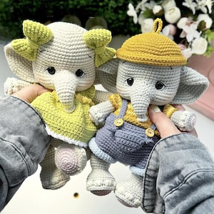 Puede incluir: Dos muñecos de elefante hechos a mano con ganchillo. Uno lleva un vestido amarillo con ribete blanco y un lazo verde. El otro lleva una camisa amarilla y un peto azul con botones amarillos y un sombrero amarillo. Los muñecos son grises, blancos, amarillos y azules.