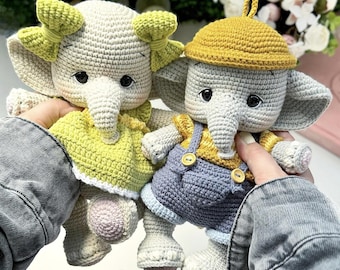 Puede incluir: Dos muñecos de elefante hechos a mano con ganchillo. Uno lleva un vestido amarillo con ribete blanco y un lazo verde. El otro lleva una camisa amarilla y un peto azul con botones amarillos y un sombrero amarillo. Los muñecos son grises, blancos, amarillos y azules.