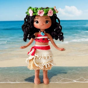 Op de afbeelding: Gehaakte pop met bruin krullend haar, die een bloemenkroon, een rood-wit topje, een witte rok met bruine franjes en een blauwe ketting draagt. De pop staat op een zandstrand.
