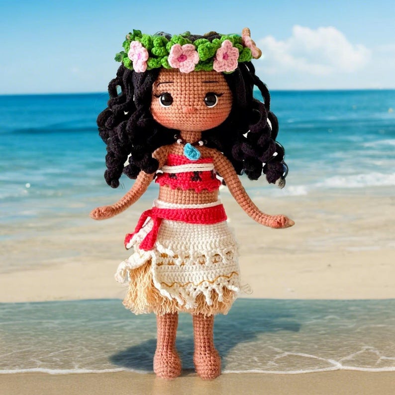 Op de afbeelding: Gehaakte pop met bruin krullend haar, die een bloemenkroon, een rood-wit topje, een witte rok met bruine franjes en een blauwe ketting draagt. De pop staat op een zandstrand.
