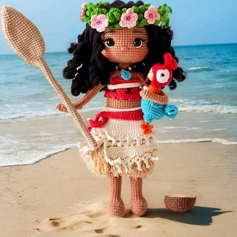 Op de afbeelding: Een gehaakte pop met bruin haar, die een bloemenkrans draagt, een rood-wit topje en een witte rok met bruine franjes. De pop houdt een blauw-rode gehaakte vogel vast. De pop staat op een zandstrand.