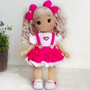May include: A handmade crochet doll with blonde curly hair and pink bows. The doll wears a white top with a pink heart, a pink skirt, and white shoes. The doll's arms and legs are beige, and it has black eyes.