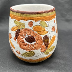 May include: A ceramic tumbler with a white base, adorned with a floral design in shades of orange, brown, and green. The rim features a brown and yellow band. The tumbler has a textured surface.