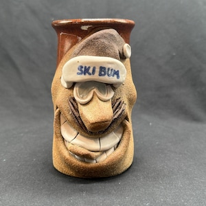 Könnte beinhalten: Keramikbecher mit einem skurrilen Gesichtsdesign. Der Becher ist hauptsächlich braun mit einem lächelnden Gesicht, Skibrille und einem Hut mit dem blauen Schriftzug "SKI BUM". Zylindrische Form.