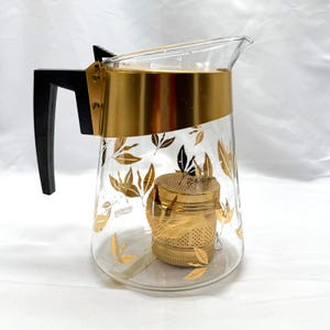 May include: A clear glass teapot featuring a gold-toned metal band around the top, a black plastic handle, and a gold-toned metal tea infuser inside. The exterior of the glass body is decorated with a repeating pattern of gold-colored leaf motifs.