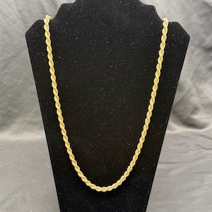 Puede incluir: Un collar de cadena de cuerda de color dorado exhibido en un expositor de joyas negro. La cadena tiene un diseño retorcido, y la estética general es clásica y elegante. El collar es una joya.