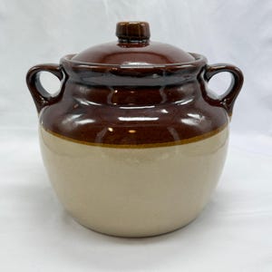 Può includere: Un vaso per fagioli in ceramica a due manici con coperchio e bordo superiore smaltati in marrone scuro lucido, che sfuma verso una base beige opaca e liscia. Il recipiente rotondo presenta due manici laterali curvi e un pomello centrale sul coperchio.