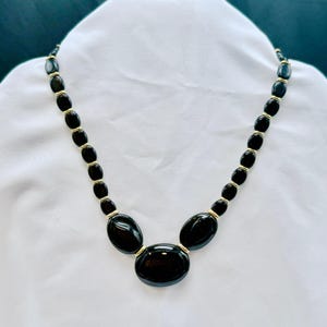 Puede incluir: Un collar de cuentas negras con detalles dorados. El collar presenta cuentas negras de forma ovalada de varios tamaños, conectadas por metal dorado. El collar se muestra sobre una superficie blanca.