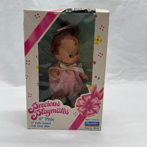 May include: A vintage 6-inch Pixie doll in its original box. The doll has red hair and is wearing a pink polka-dot dress. The box features the "Precious Playmates" logo and the text "6" Fully Jointed Soft Vinyl Skin."
