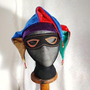 Peut inclure: Un chapeau de bouffon multicolore avec un masque assorti. Le chapeau est fait de tissu bleu, rouge, vert et or avec une bande violette. Le masque noir a une bordure orange. L'ensemble est présenté sur une tête de mannequin noire.