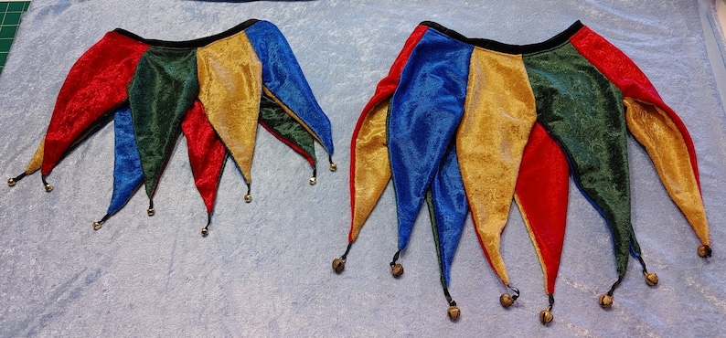 K&ouml;nnte beinhalten: Zwei Narrenkappen mit Gl&ouml;ckchen. Die Kappen sind aus rotem, gr&uuml;nem, blauem und goldenem Stoff. Die Kappen haben ein schwarzes Band um die Oberseite.