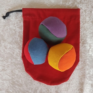 May include: Three multi-colored fabric juggling balls resting on a solid red drawstring pouch. The balls feature panels of purple, dark green, blue, red, yellow, and orange fabric. The red pouch has a black drawstring cord with a knotted end.