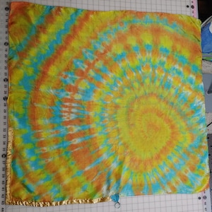 May include: A square, tie-dye textile with a spiral pattern in shades of yellow, orange, and blue. The fabric has a gold-colored trim along the edges. The textile measures approximately 44 inches in length and width.