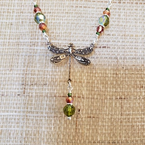 May include: Silver-toned dragonfly pendant necklace with a delicate chain. The dragonfly has intricate wing detailing. The necklace features green, orange, and iridescent beads. The pendant hangs from a silver chain, creating a nature-inspired jewelry piece.