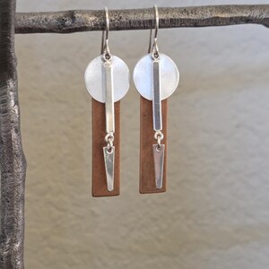 May include: A pair of silver and copper-toned earrings. Each earring features a silver disc, a vertical silver bar, a copper-colored rectangular shape, and a silver, inverted triangle pendant. The earrings hang from silver hooks.
