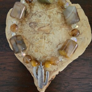May include: A beaded bracelet featuring a silver cactus charm. The bracelet is made with a mix of brown and tan-colored stones and clear beads. The bracelet is displayed on a light brown, textured surface.