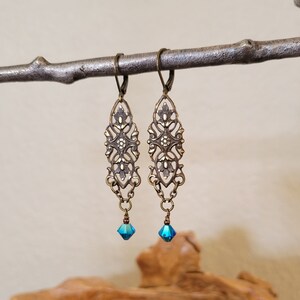 May include: Ornate bronze-toned earrings with an open filigree design. Each earring features a dangling, faceted, teal-colored crystal. The earrings are suspended from a metal branch against a neutral background.