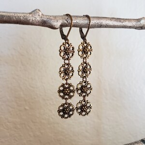 May include: A pair of antique bronze-colored earrings. Each earring features a series of four openwork floral designs connected by small links. The earrings hang from a lever-back closure. The earrings are displayed on a silver-colored branch.