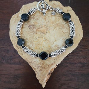 May include: A black and silver beaded bracelet with a toggle clasp. The bracelet features black square beads and silver beads with a textured surface.