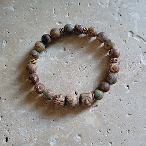 May include: A beaded bracelet featuring a mix of brown, green, and tan stone beads. The bracelet includes several square-shaped beads with carved details and copper-colored spacers. The bracelet is designed to be worn around the wrist.