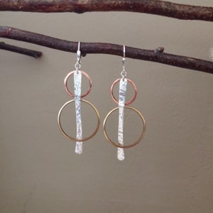 May include: A pair of modern earrings with a mixed metal design. Each earring features a silver hammered bar suspended between a copper ring and a gold-colored ring. The earrings hang from silver ear wires.