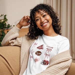May include: White long-sleeve t-shirt with maroon football-themed graphics, including a football, helmet, bows, and a megaphone with the word "fabulous". The shirt is decorated with watercolor-style illustrations.