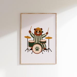 Può includere: Una stampa artistica da parete incorniciata raffigurante un orso bruno con un berretto giallo e occhiali da sole, che indossa una camicia a righe verdi e bianche. L'orso suona la batteria con due bacchette e la grancassa ha un'impronta di zampa.
