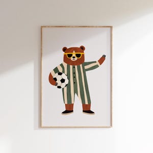 Op de afbeelding: Een ingelijste kunstdruk met een cartoonbeer met een gele zonnebril en een groen en wit gestreept pak. De beer houdt een zwart-witte voetbal vast en zwaait. Het kunstwerk heeft een speelse, grillige stijl.