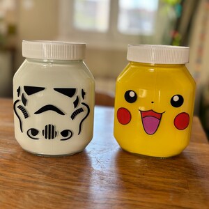 Peut inclure: Deux bocaux décoratifs, l'un blanc avec un motif de Stormtrooper noir et l'autre jaune avec un visage de Pikachu. Les deux bocaux ont des couvercles blancs et sont posés sur une surface en bois.