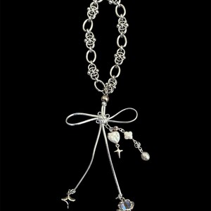 May include: A silver-toned necklace with a delicate chain and a pendant featuring a bow, heart, star, and other charms. The necklace has a polished finish and is set against a black background.