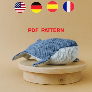 Puede incluir: Un juguete de ballena azul y gris tejido a ganchillo con el texto "PDF PATTERN" en rojo. La ballena está sobre un pequeño soporte redondo de madera. La ballena tiene una barriga gris claro y un cuerpo azul oscuro.