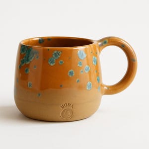May include: A ceramic mug with a rounded shape and a large handle. The mug has a two-tone design, with a tan base and a warm orange top, speckled with turquoise accents. The word "MORA" is stamped on the base.
