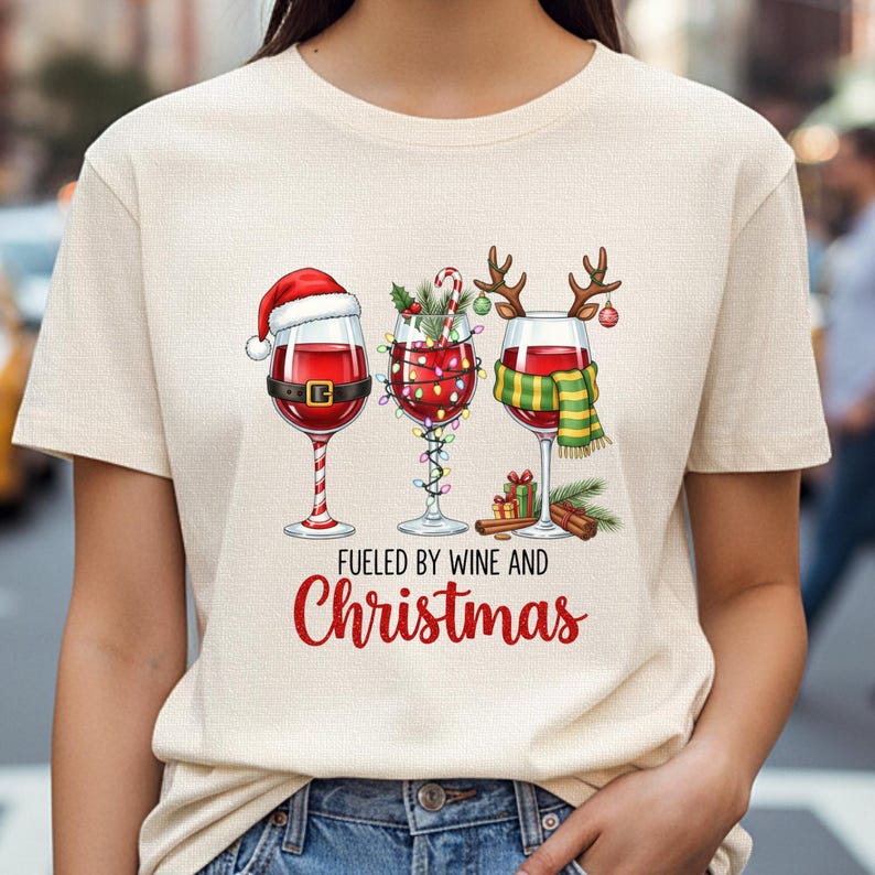 Peut inclure: T-shirt beige avec trois verres &agrave; vin d&eacute;cor&eacute;s pour No&euml;l. Un verre porte un bonnet de P&egrave;re No&euml;l, un autre est orn&eacute; de guirlandes lumineuses, et le troisi&egrave;me a des bois de renne. Le texte "Fueled by Wine and Christmas" est en dessous.