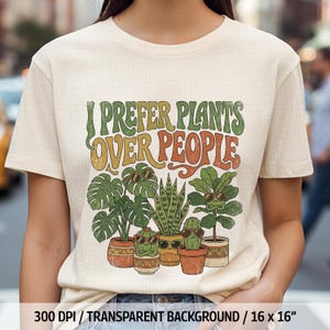 May include: Cream-colored t-shirt featuring the text "I Prefer Plants Over People" in green and orange retro font. Below the text are illustrations of potted plants wearing sunglasses. The shirt has a crew neck and short sleeves.