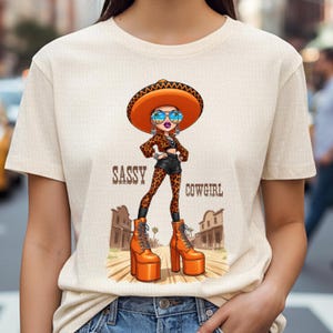 May include: A beige t-shirt featuring a cartoon cowgirl with a sombrero, sunglasses, and platform boots. The cowgirl is wearing a leopard print outfit. The text "SASSY COWGIRL" is printed on the shirt.