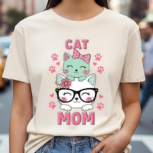 Könnte beinhalten: Beiges T-Shirt mit einem Aufdruck von zwei Cartoon-Katzen. Die obere Katze ist mintgrün mit einer rosa Schleife, die untere Katze ist weiß mit Brille. Die Worte "CAT" und "MOM" sind aufgedruckt.