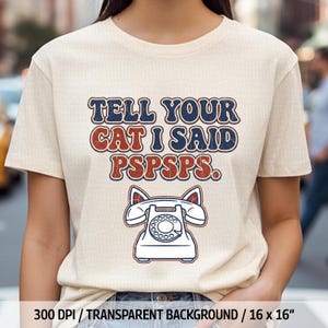 以下が含まれることがあります： ベージュ色のTシャツ。テキスト「TELL YOUR CAT I SAID PSPSPS.」が青と赤のレトロなフォントでプリントされています。テキストの下には、猫の耳が付いた白い電話のイラスト。Tシャツの幅は40.6cmです。