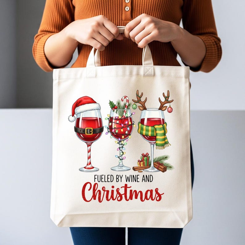 Peut inclure: Un sac fourre-tout cr&egrave;me avec trois verres &agrave; vin d&eacute;cor&eacute;s pour No&euml;l. Les verres sont remplis de vin rouge et orn&eacute;s d'un bonnet de P&egrave;re No&euml;l, de guirlandes lumineuses et de bois de rennes. Le texte "Fueled by Wine and Christmas" est imprim&eacute; en rouge.
